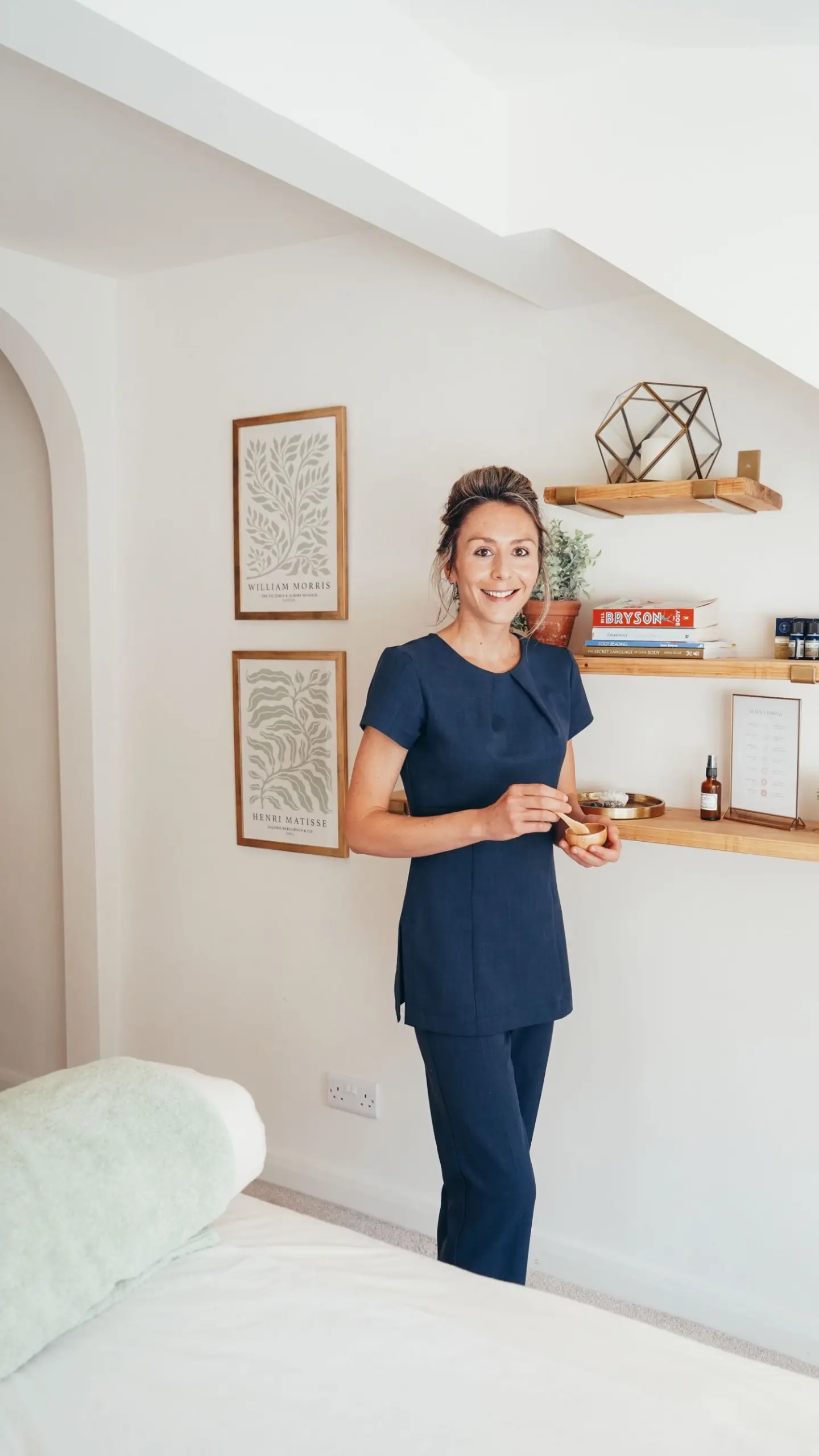 Ellie holding reflexology equipment while standing in front of posters and wooden shelves in the Rutland Reflexology clinic treatment room